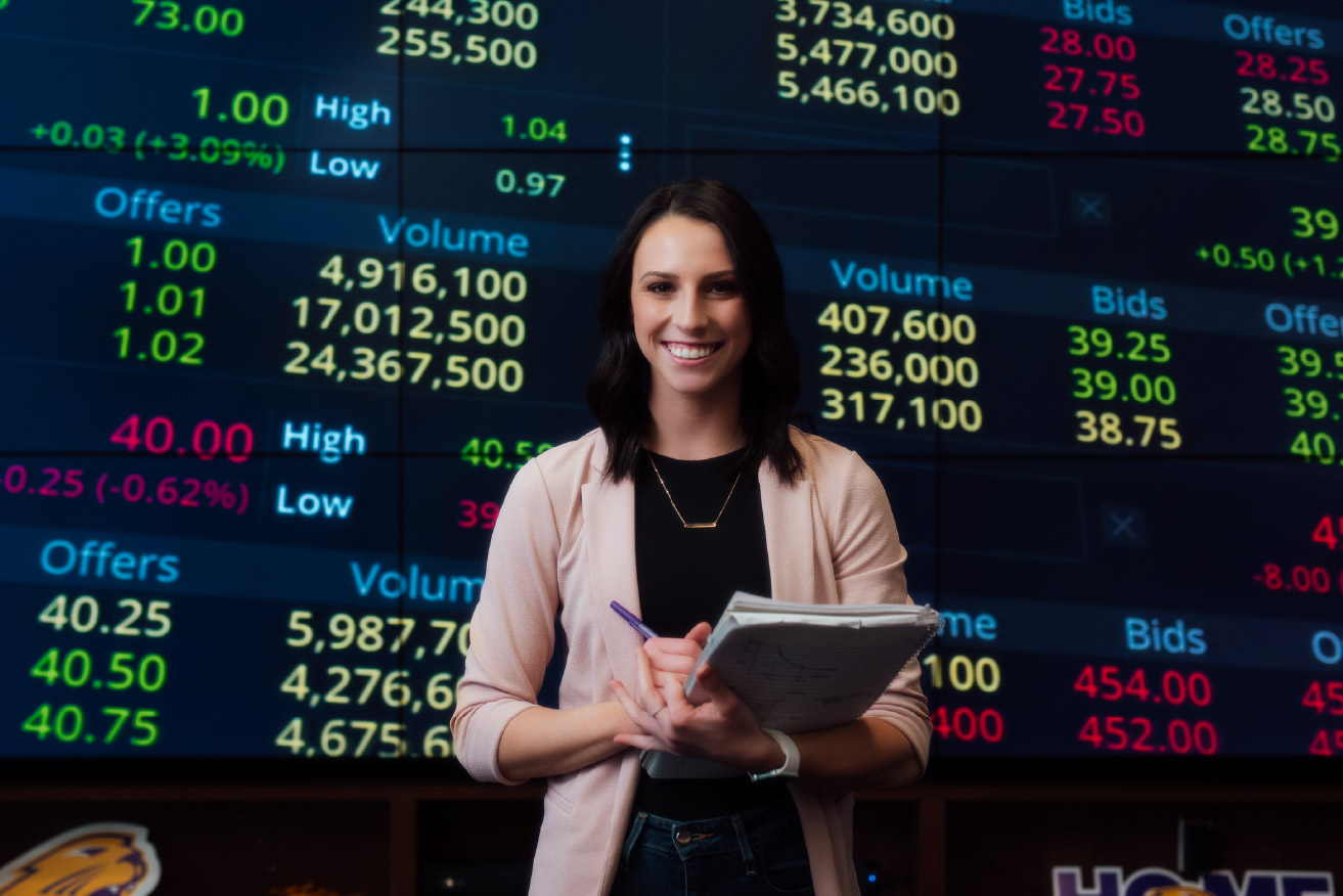 Student holding a notebook while standing in front of a large screen displaying stock market data.