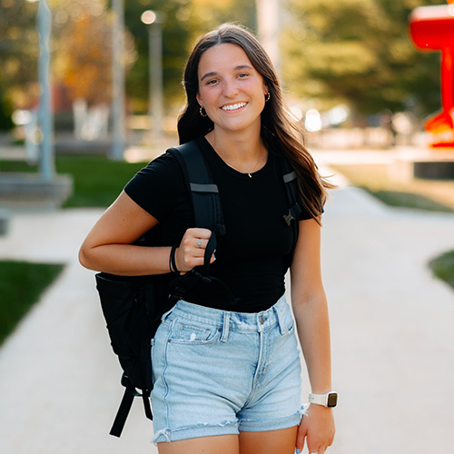 Smiling student pursuing a business degree walking on campus with a backpack on a sunny day.